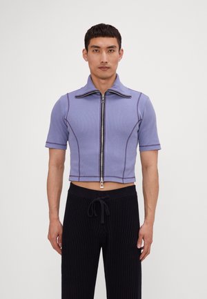 Hellpurpurnes, kurz geschnittenes Zip-Up-Shirt mit kurzen Ärmeln, gerippter Textur, kontrastierenden Nähten und hohem Kragen. Kombiniert mit schwarzen, strukturierten Hosen.