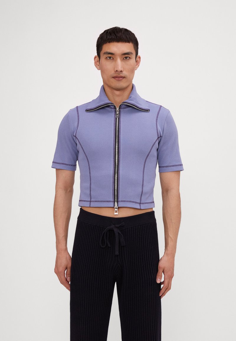 Hellpurpurnes, kurz geschnittenes Zip-Up-Shirt mit kurzen Ärmeln, gerippter Textur, kontrastierenden Nähten und hohem Kragen. Kombiniert mit schwarzen, strukturierten Hosen.