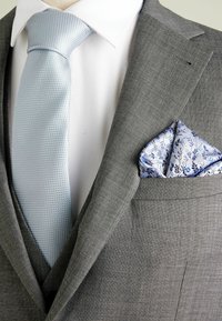 Veste de costume grise avec une cravate texturée bleu clair et un mouchoir de poche à motifs présentant des motifs floraux bleus sur fond blanc.
