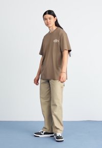 Brun kortermet T-skjorte med en liten logo, kombinert med beige bukser og svarte joggesko. Innstillingen har en enkel lyse bakgrunn.