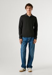 Černá polokošile s dlouhým rukávem a límečkem, zkombinovaná se světle modrými rovnými džíny a bílými teniskami s černými akcenty. Jednoduché pozadí.