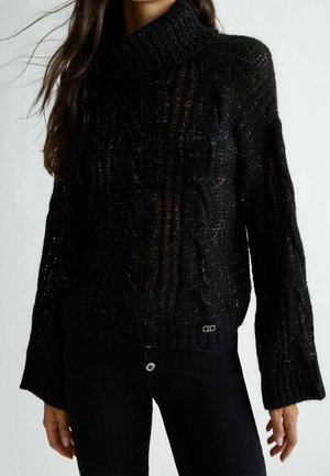 Femme portant un pull col roulé noir, tricoté de manière lâche, avec des manches longues et un pantalon noir ajusté sur un fond uni.