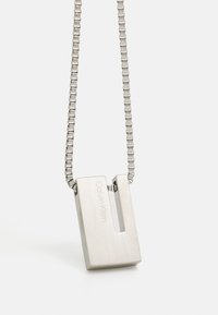 Collar de pendant de acero inoxidable con un diseño rectangular, que presenta un recorte y grabado "Calvin Klein." Unido a una cadena texturizada.