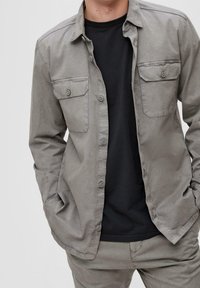 Grijs overhemd van stof met een gladde textuur, met twee borstzakken en een kraag. Draagt over een zwart shirt.