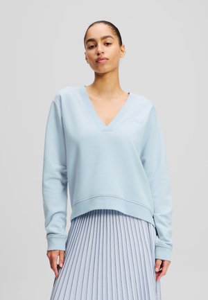 Light blue V-neck sweatshirt with a relaxed fit, ribbed cuffs, and a subtle embroidered logo. Paired with a pleated, light blue skirt.