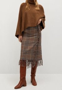 Poncho lavorato a maglia marrone, gonna plaid avvolgente con orlo frangiato e stivali al ginocchio in pelle marrone. I tessuti sono morbidi e dalla texture interessante.