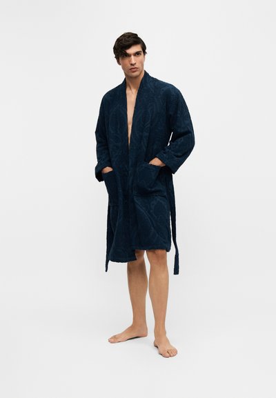 Homme debout pieds nus portant un peignoir texturé bleu foncé, les mains dans les poches, sur un fond clair uni.