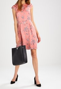 Rosa Blumenkleid mit kurzen Kappenärmeln, ausgestellter Silhouette, schwarzer Handtasche mit Metallakzenten und schwarzen hochhackigen Schuhen.