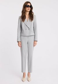 Jasnoszara marynarka dopasowana z podwójnym zapięciem, w zestawie z dopasowanymi spodniami o slim fit. Modelka nosi duże czarne okulary przeciwsłoneczne oraz beżowe szpilki.