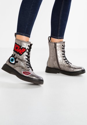 Bottines argentées métalliques à lacets avec fermeture éclair, décorées d'un patch rouge « POW ! », un œil bleu et des lèvres rouges, portées avec un jean skinny foncé.