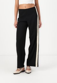 Pantalons larges noirs avec une rayure crème sur le côté, présentant une texture lisse et une coupe décontractée. Assortis à des chaussures à brides beiges.