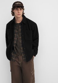 Sort fleecetrøje med to forlommer over en ternet skjorte. Brune cargobukser med sidelommer. Brun kasket med broderet tekst.