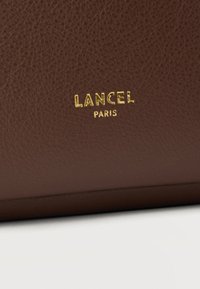 Καφέ δερμάτινη υφή με ανάγλυφο χρυσό λογότυπο "LANCEL PARIS" στο κέντρο.