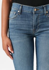 Fitted blue denim jeans with a mid-rise waist, five-pocket design, and gold stitching details, showcasing a smooth, slightly faded texture.