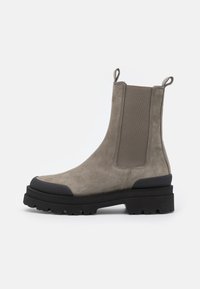 Taupe Wildleder Chelsea-Boot mit schwarzem groben Profilsohle, elastischen Seitenpanelen und Zuglaschen an den Knöcheln vor weißem Hintergrund.