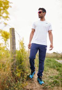 T-shirt bianco con collo a giro, pantaloni sportivi blu navy e scarpe da trekking turchesi. In piedi su un sentiero erboso con fiori gialli nelle vicinanze.