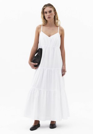 Kobieta ubrana w białą, warstwową suknię maxi na cienkich ramiączkach, trzymająca czarną kopertówkę i nosząca czarne płaskie buty, stojąca na tle białego tła.