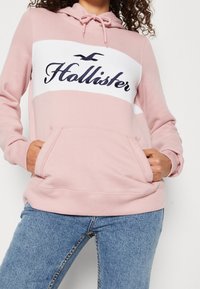 Žena v růžové mikině s bílým panelem a textem "Hollister", s rukama v přední kapse, kombinováno s modrými džíny.