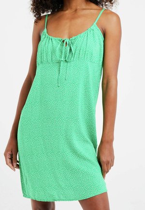 Kvinde iført en grøn ærmeløs kjole med hvide prikker og en knude foran ved halsudskæringen mod en hvid baggrund.