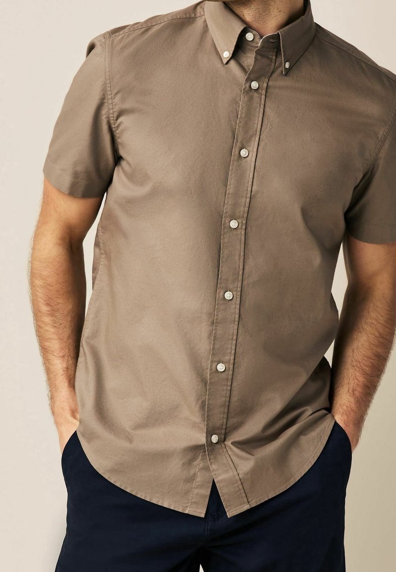 Homme portant une chemise beige à manches courtes avec col, mains dans les poches, associée à un pantalon bleu marine foncé.