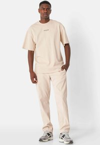Camiseta de manga corta beige con un pequeño logo en el pecho, combinada con pantalones beige a juego y zapatillas grises con detalles en negro.