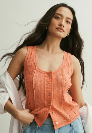 Junge Frau mit langen dunklen Haaren, die ein ärmelloses orangefarbenes, geknöpftes Top und hellblaue Jeans trägt, hält ein weißes Hemd über den Armen.