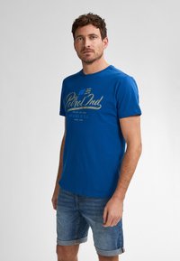 Modré bavlněné tričko s krátkým rukávem, se zlatým potiskem loga "Petrol Ind". V kombinaci s džínovými šortkami, které předvádějí ležérní styl.