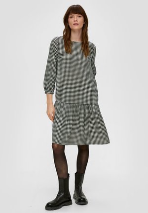 Czarna i biała sukienka w wzór houndstooth z rękawami do trzech czwartych, dopasowanym korpusem i haftowanym, rozkloszowanym dołem. Stylizowana z czarnymi botkami za kostkę.