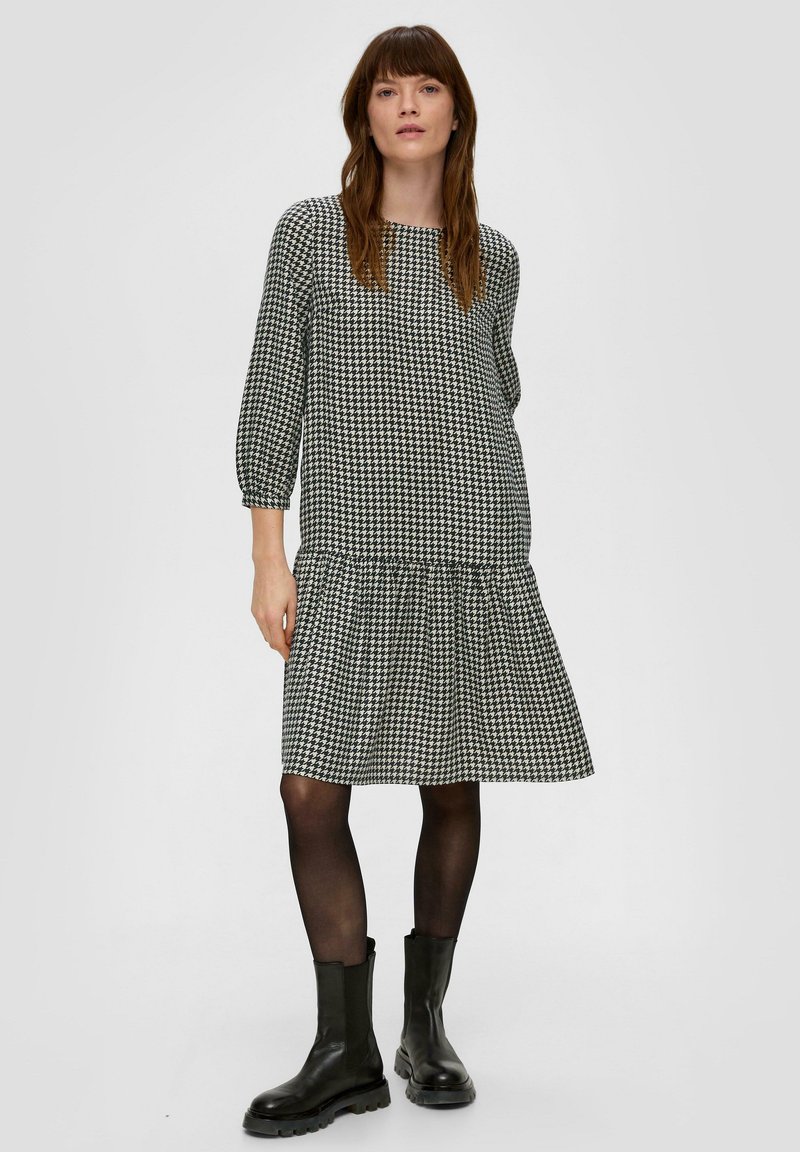 Czarna i biała sukienka w wzór houndstooth z rękawami do trzech czwartych, dopasowanym korpusem i haftowanym, rozkloszowanym dołem. Stylizowana z czarnymi botkami za kostkę.