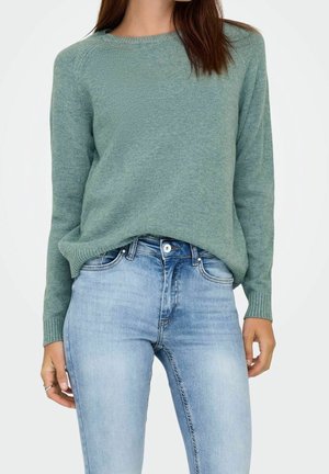 Jeune femme portant un pull vert clair et un jean taille haute bleu clair, la main reposant le long du corps.
