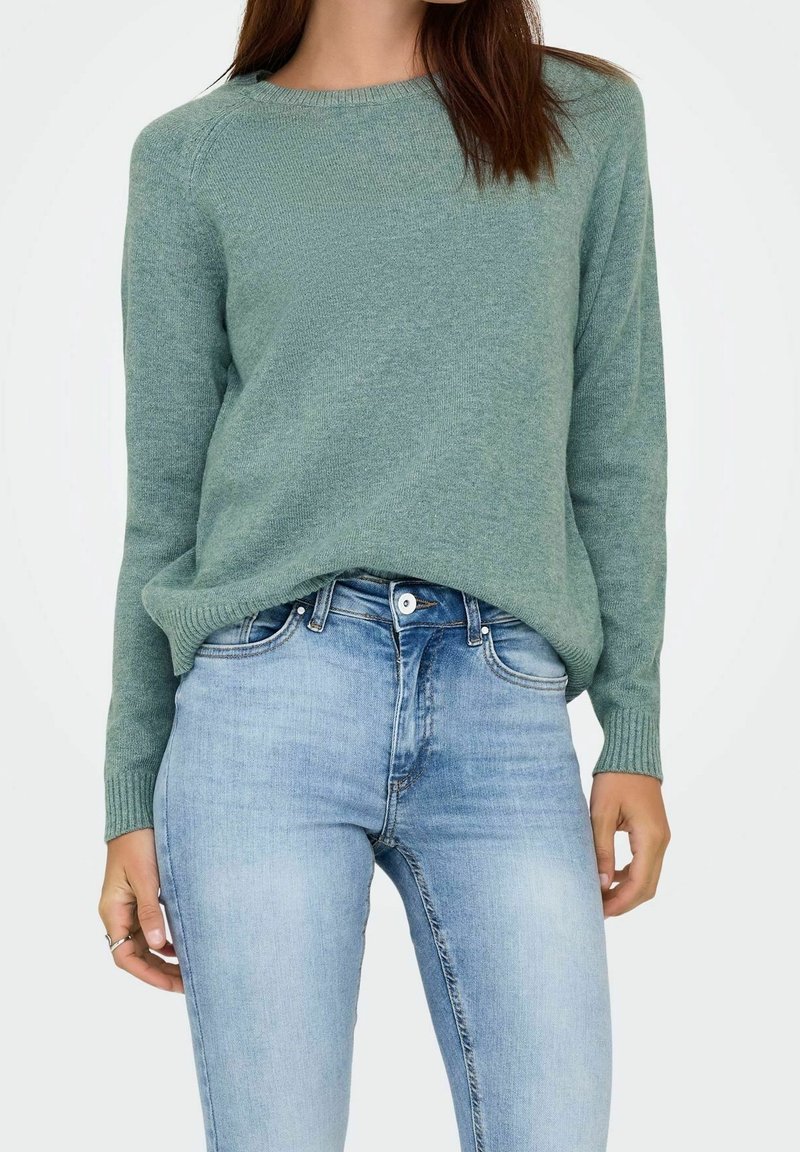 Jeune femme portant un pull vert clair et un jean taille haute bleu clair, la main reposant le long du corps.