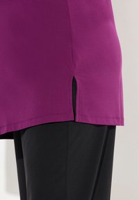 Tunic viola con una texture liscia, caratterizzata da aperture laterali e un orlo dritto, abbinata a pantaloni neri. Design semplice, senza motivi visibili.