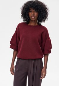 Bordowy sweter z dzianiny z krótkimi, szerokimi rękawami i okrągłym dekoltem, zestawiony z ciemnobrązowymi, wysokim stanem spodniami. Gładka tekstura.