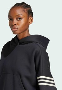 Sudadera negra con capucha y bolsillo frontal, con detalles de franjas blancas en las mangas. Tejido suave, ajuste casual.