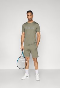 Αθλητική στολή ελιάς με μπλουζάκι λαιμού με το λογότυπο "BOSS", ταιριαστά σορτς και ρακέτα τένις, σε συνδυασμό με λευκά αθλητικά παπούτσια.