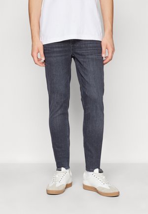 Man draagt slim-fit donkergrijze jeans, wit T-shirt en witte sneakers met rubberen zolen, staand tegen een effen lichte achtergrond.