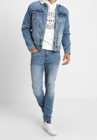 Giacca di jeans con colletto in sherpa, azzurro chiaro, tasche frontali. Felpa bianca con logo rotondo, jeans azzurri chiari e sneakers bianche.