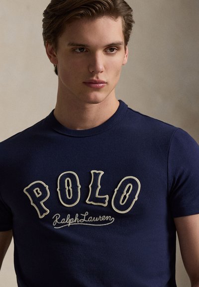Μπλε μπλουζάκι από βαμβάκι με ανάγλυφο λογότυπο "POLO" σε εκρού χρώμα και κεντημένο το κείμενο "Ralph Lauren" από κάτω. Σύνθεση με κοντά μανίκια και στρογγυλή λαιμόκοψη.