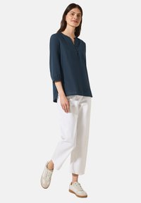 Blusa blu navy a maniche lunghe con chiusura a bottoni, abbinata a pantaloni bianchi larghi e sneakers chiare.
