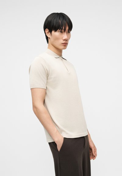 Jeune homme aux cheveux noirs portant un polo beige à manches courtes et un pantalon foncé, debout les mains dans les poches devant un fond uni.