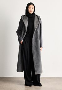 Dlhý sivý vonkajší kabát s jemnou textúrou, veľkou kapucňou a širokými klopami, nosený cez čierne tričko a voľné čierne nohavice.