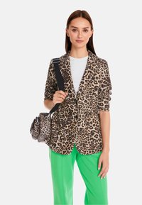 Leopard print blazer in soft fabric, single-breasted design, two front pockets. Worn with a white shirt and bright green pants, holding a bag.