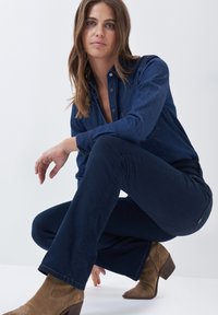 Mørk denimskjorte knappet ved kraven, parret med marineblå flared jeans. Lysebrune hælede støvler med dekorativ syning på siden.