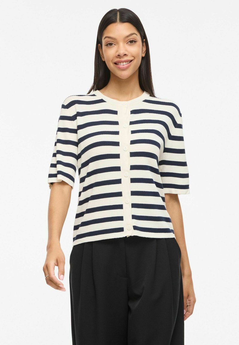 Femme souriante, portant un cardigan blanc à manches courtes avec des rayures horizontales bleu marine et un pantalon plissé noir, debout sur fond blanc.