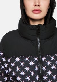 Schwarze Steppjacke mit hohem Kragen, die im unteren Bereich ein Sternenmuster in Weiß und Rot aufweist, sowie einen Vorderreißverschluss mit glänzender Oberfläche.