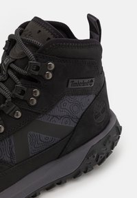 Botte de randonnée noire imperméable avec des panneaux texturés noirs et gris, des œillets en métal et une semelle robuste. Présence du logo Timberland sur le côté.