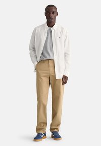 White button-up shirt over a grey t-shirt, paired with tan trousers and blue sneakers with white accents; smooth fabric, relaxed fit.