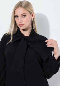 Sort bluse med en stor bundet sløjfe ved halsen, lange ærmer og knapmanchetter. Modellen bærer store sølv hoop-øreringe mod en ensfarvet baggrund.