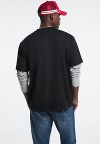 T-shirt nera a maniche corte sopra una maglietta grigia a maniche lunghe, abbinata a jeans blu e un cappellino rosso con strisce bianche.