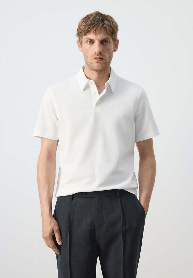 Homme aux cheveux courts portant un polo blanc à manches courtes et un pantalon foncé, debout devant un fond clair uni.